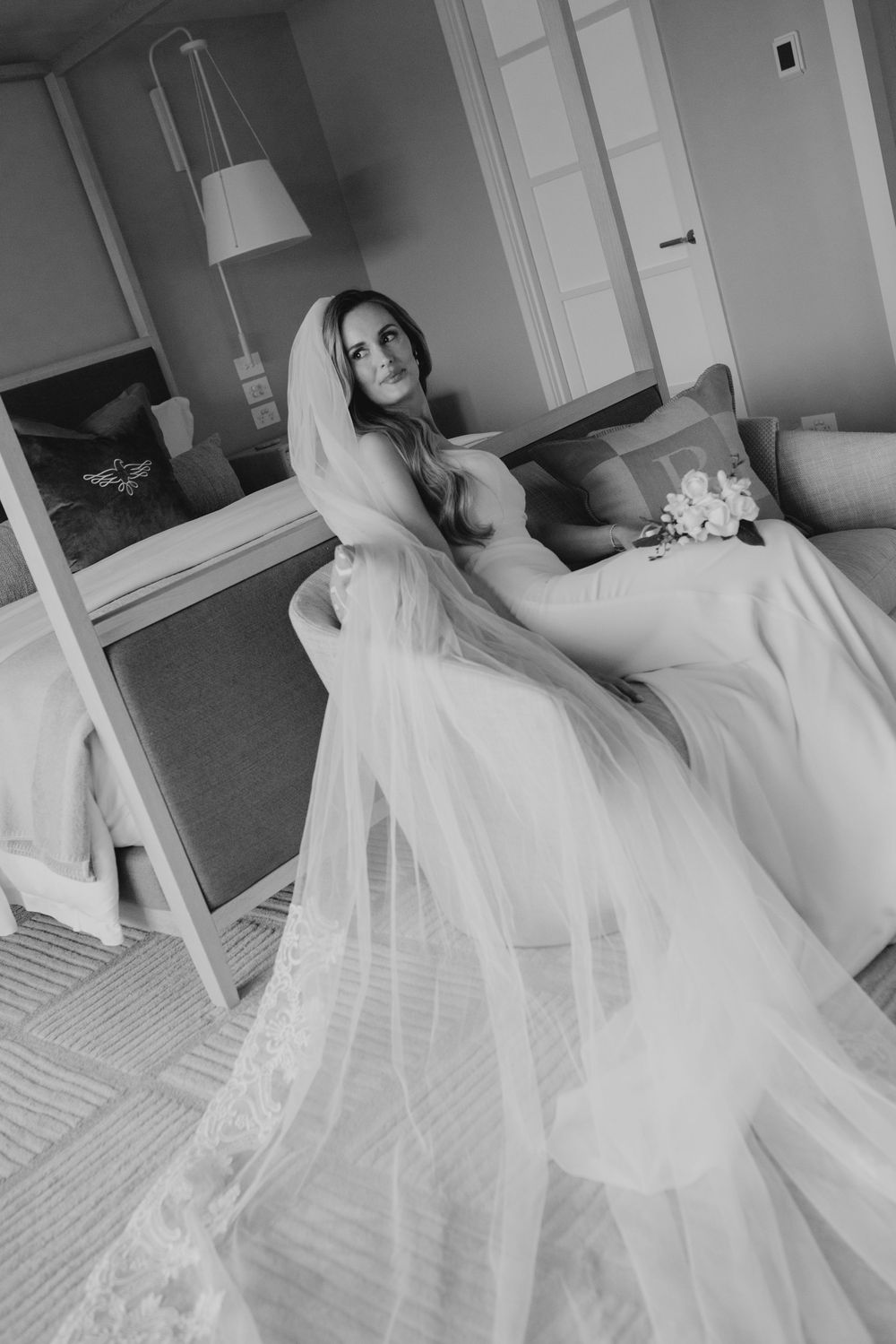Black and white photo of a bride in a flowing wedding gown and veil, seated elegantly in a hotel room.