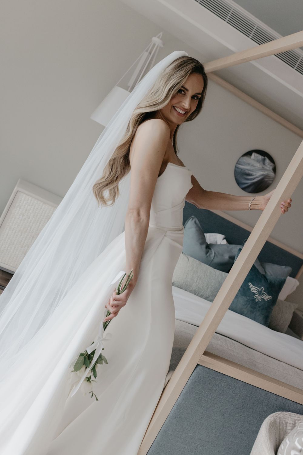 Smiling bride in white wedding gown and veil holding flowers, posing in a hotel room.