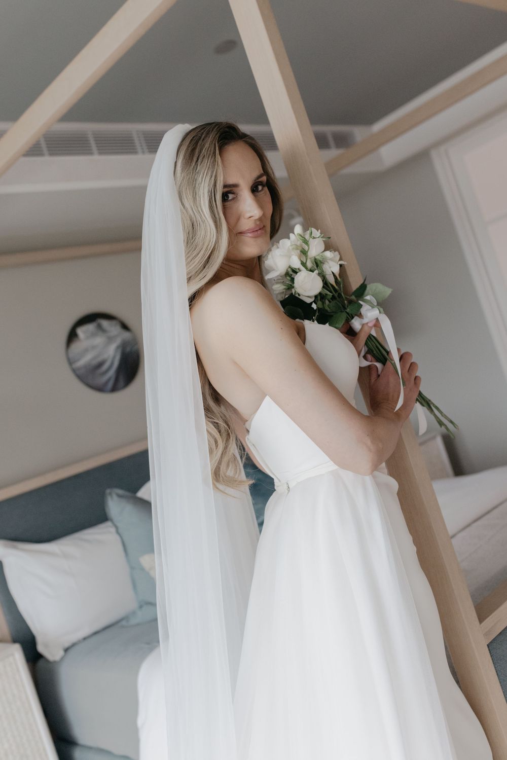Bride in strapless white gown and veil holds bouquet of white flowers, posing elegantly in a softly lit bedroom.