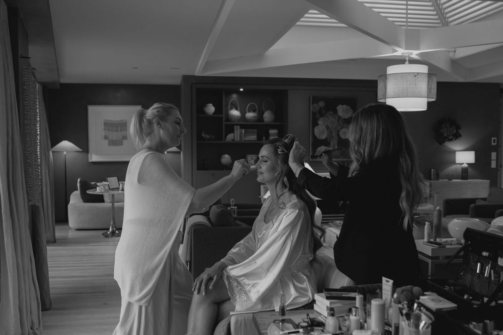 Black and white photo of a bride getting hair and makeup done by two stylists in an elegant room.