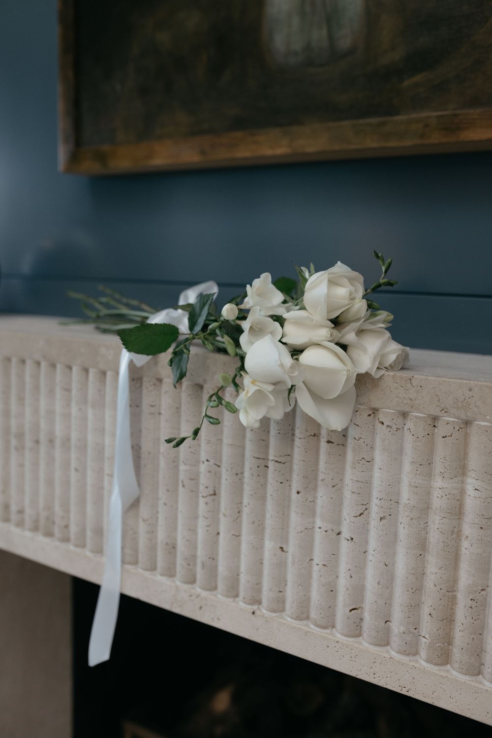 White rose bouquet with ribbon resting on a textured stone fireplace mantel against a blue wall.