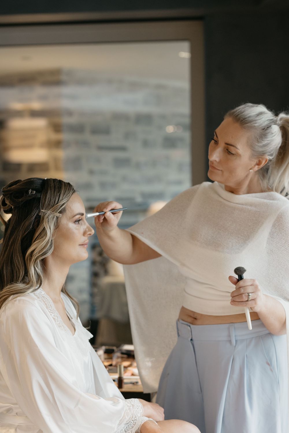 A makeup artist applying eyeshadow to a bride in a white robe during wedding preparations.