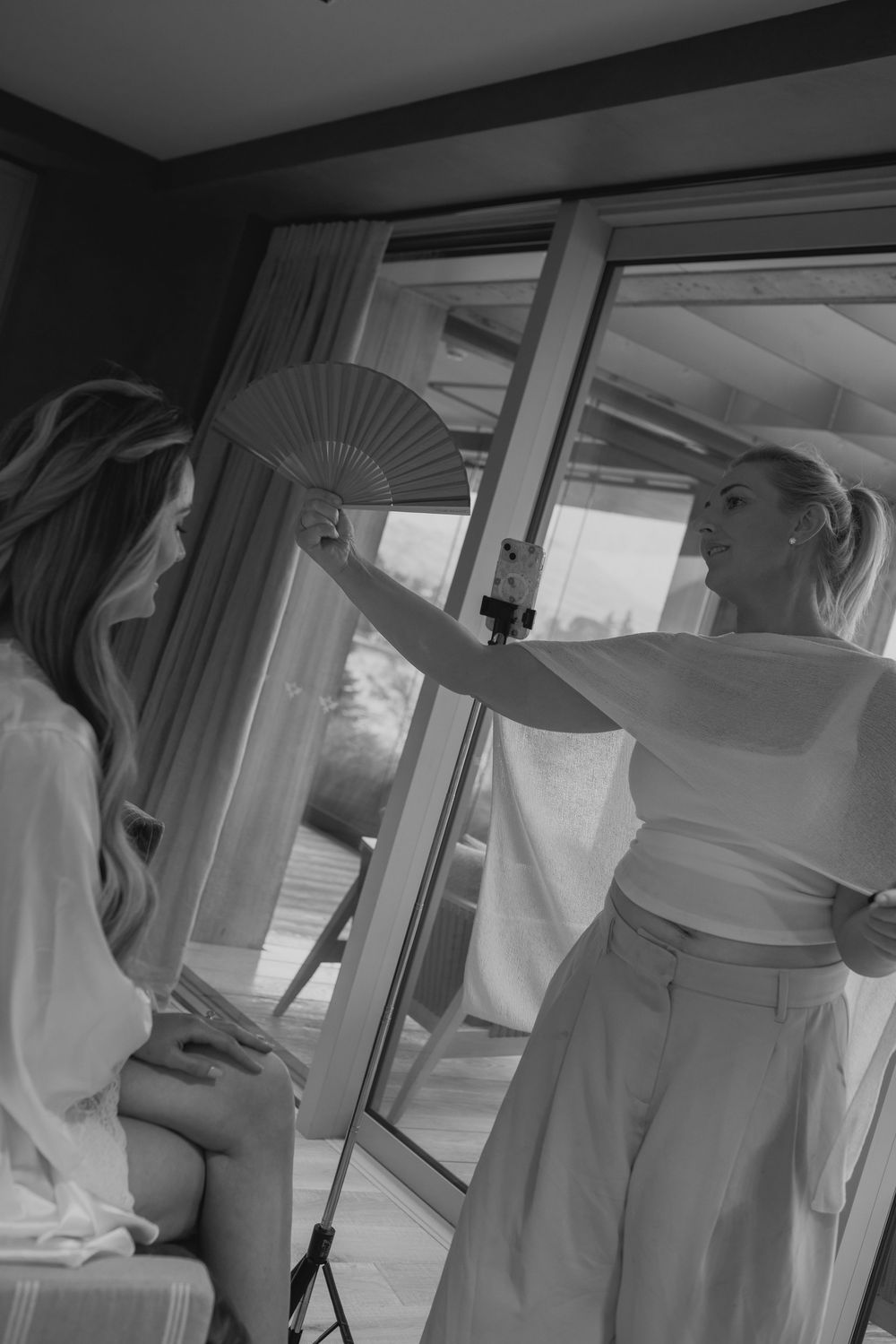 Black and white photo of two women in white outfits, one holding a decorative fan near a window.