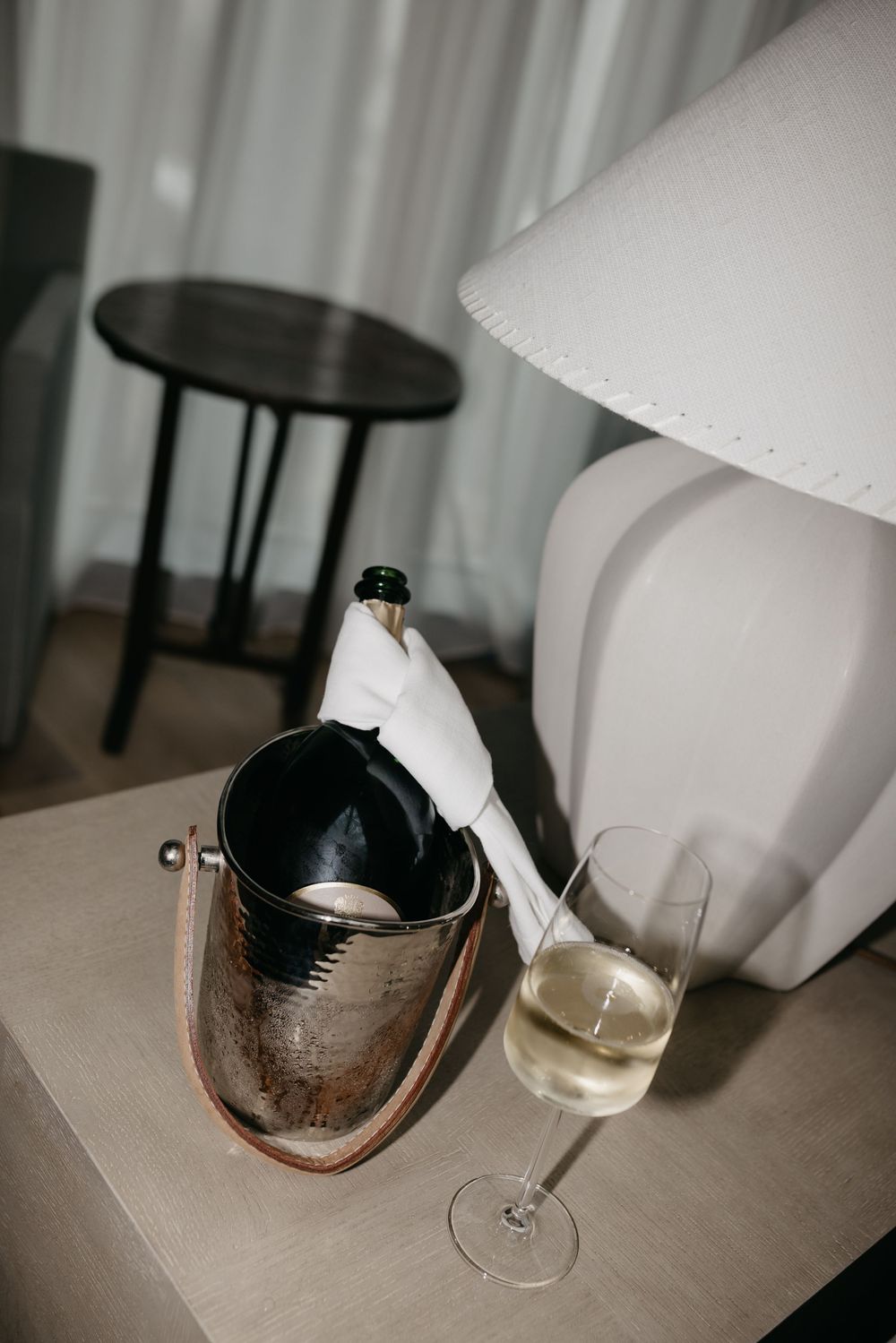 Champagne bottle in silver ice bucket beside a full flute glass on nightstand with white lamp in hotel room.
