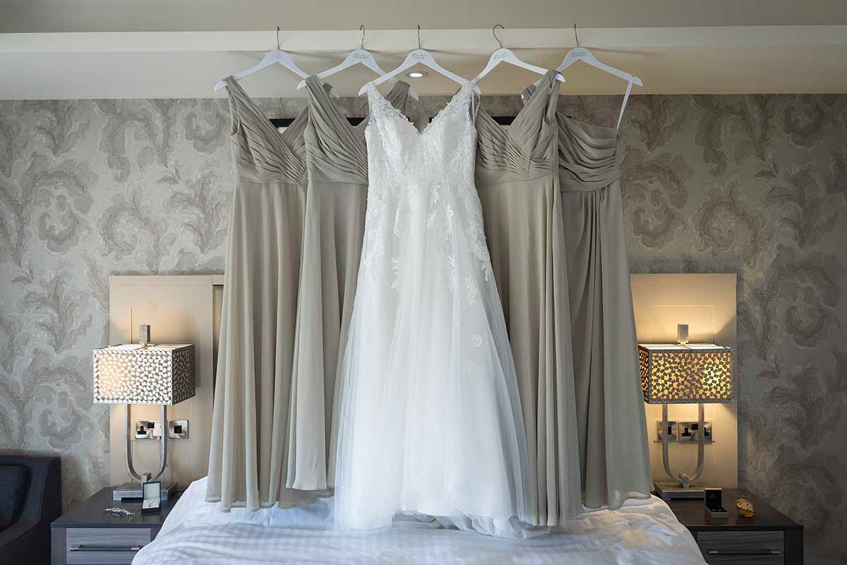 White wedding gown flanked by four silver bridesmaid dresses hanging on hangers above a bed in an elegant hotel room.