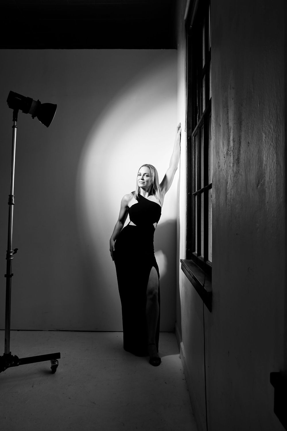 Black and white photo of a blonde woman in an elegant black gown posing near a window with studio lighting.