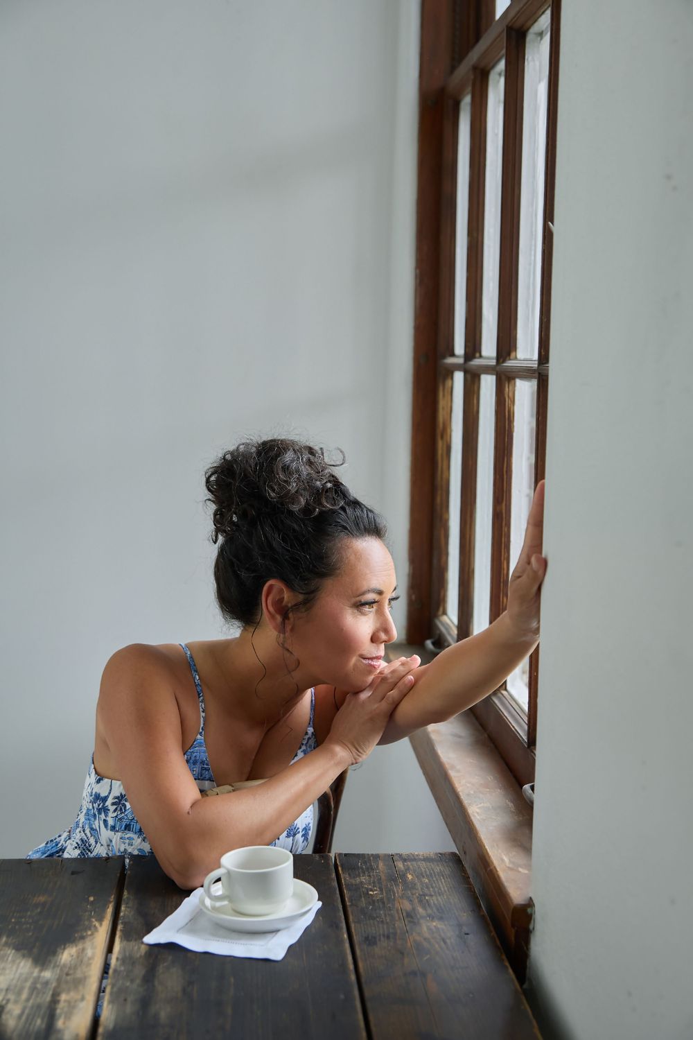 Woman in floral dress gazing out wooden window, resting chin on hands beside white coffee cup on rustic table.