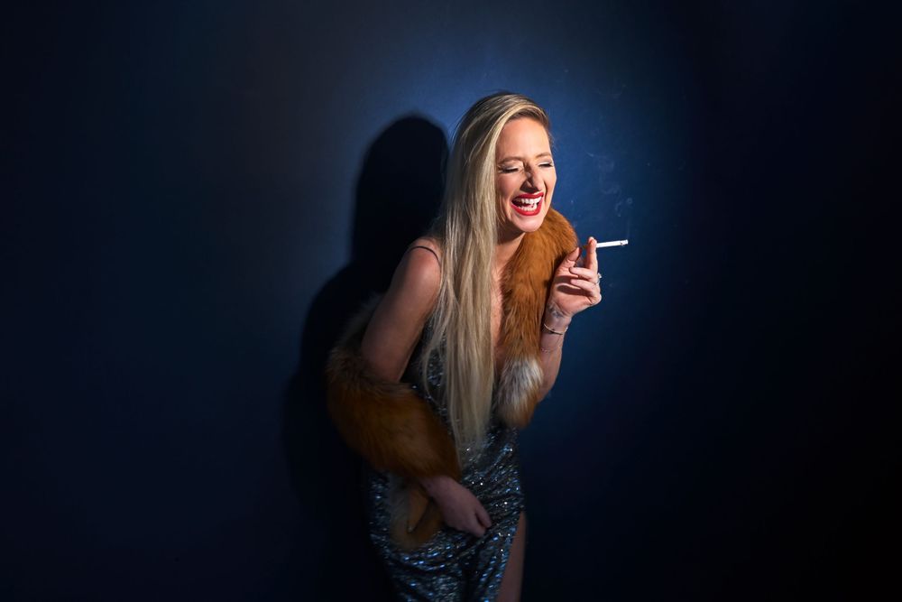 Glamorous blonde woman laughing while smoking a cigarette, wearing a fur stole and sequin dress against dark background.