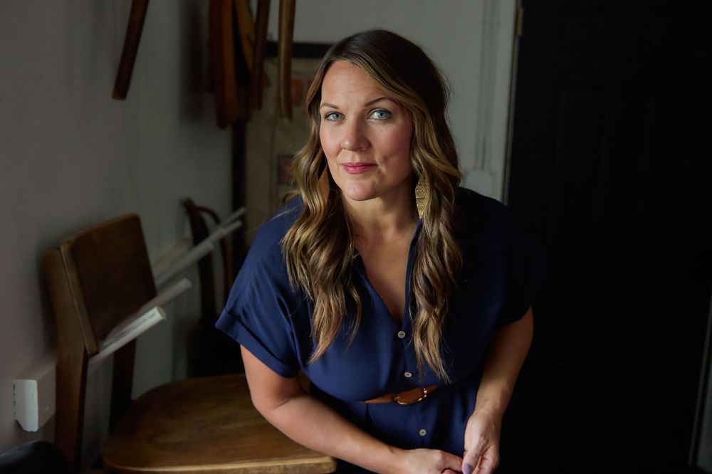 Woman in blue blouse sitting thoughtfully by a window in a softly lit indoor setting.