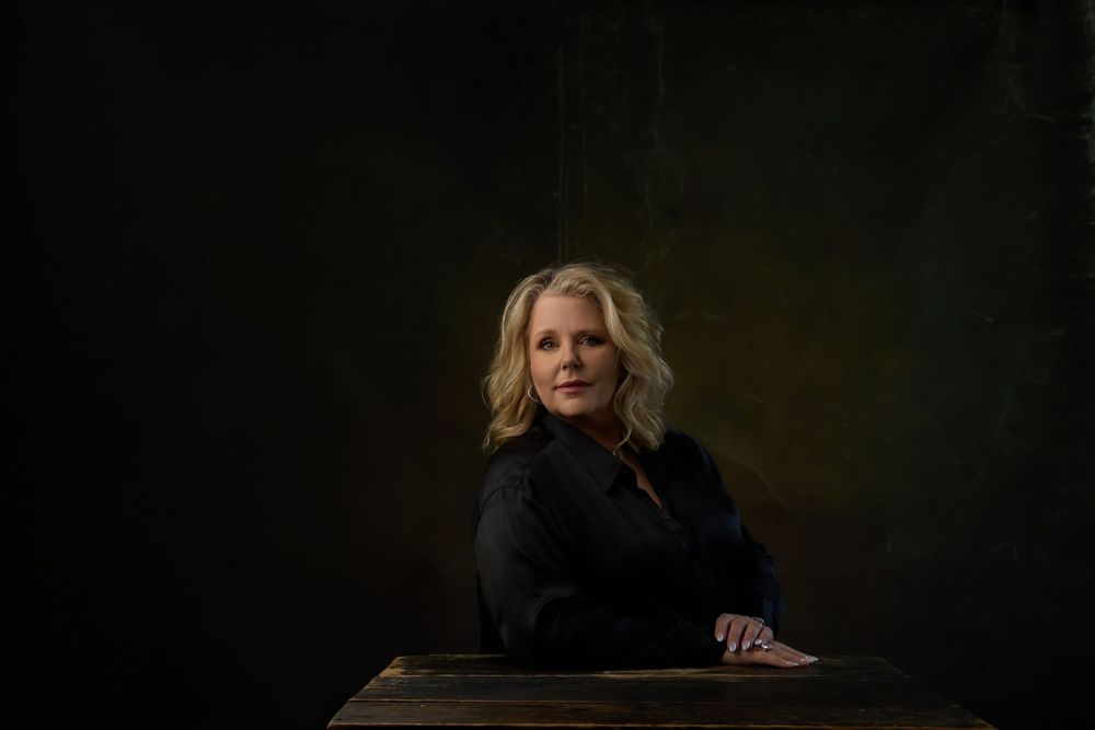 Blonde woman in black jacket poses at wooden table against dark green backdrop in professional portrait.
