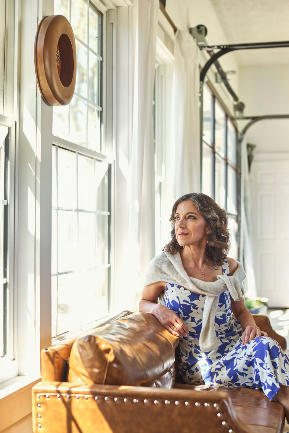 A woman in a blue floral dress sits on a leather sofa gazing out bright windows in a sunlit room.