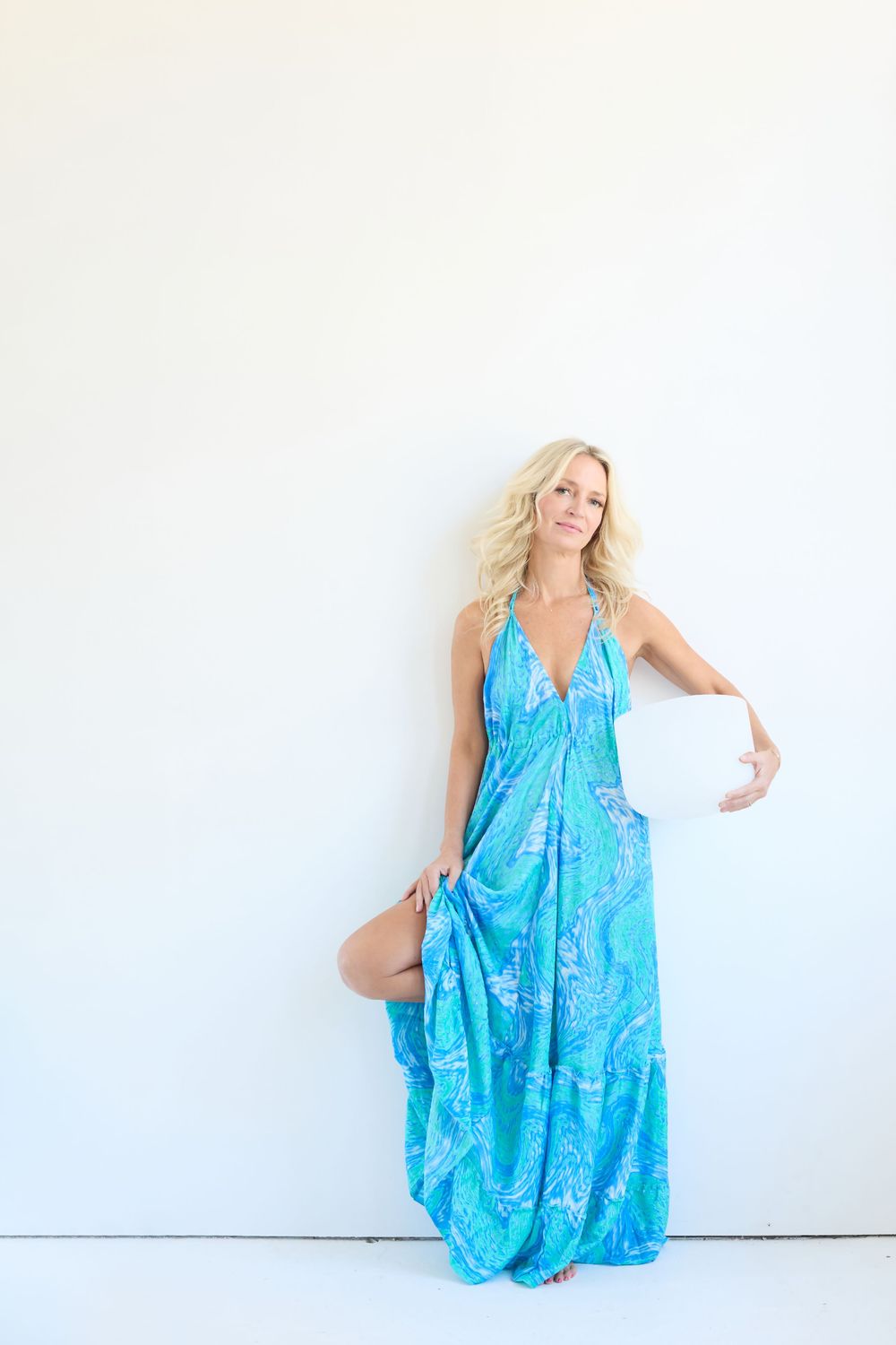 Blonde woman in a blue paisley halter maxi dress leaning against a white wall holding a white clutch.