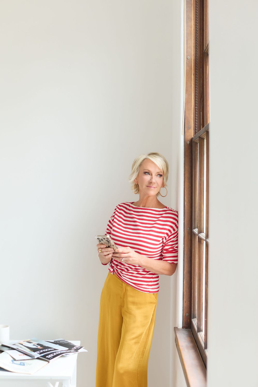 Blonde woman in red striped top and yellow pants leaning against wall holding smartphone near window.
