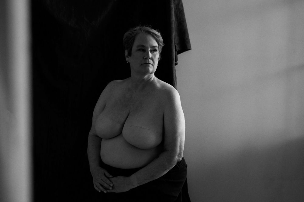 Black and white portrait of an older woman posing topless against a dark backdrop in a studio setting.