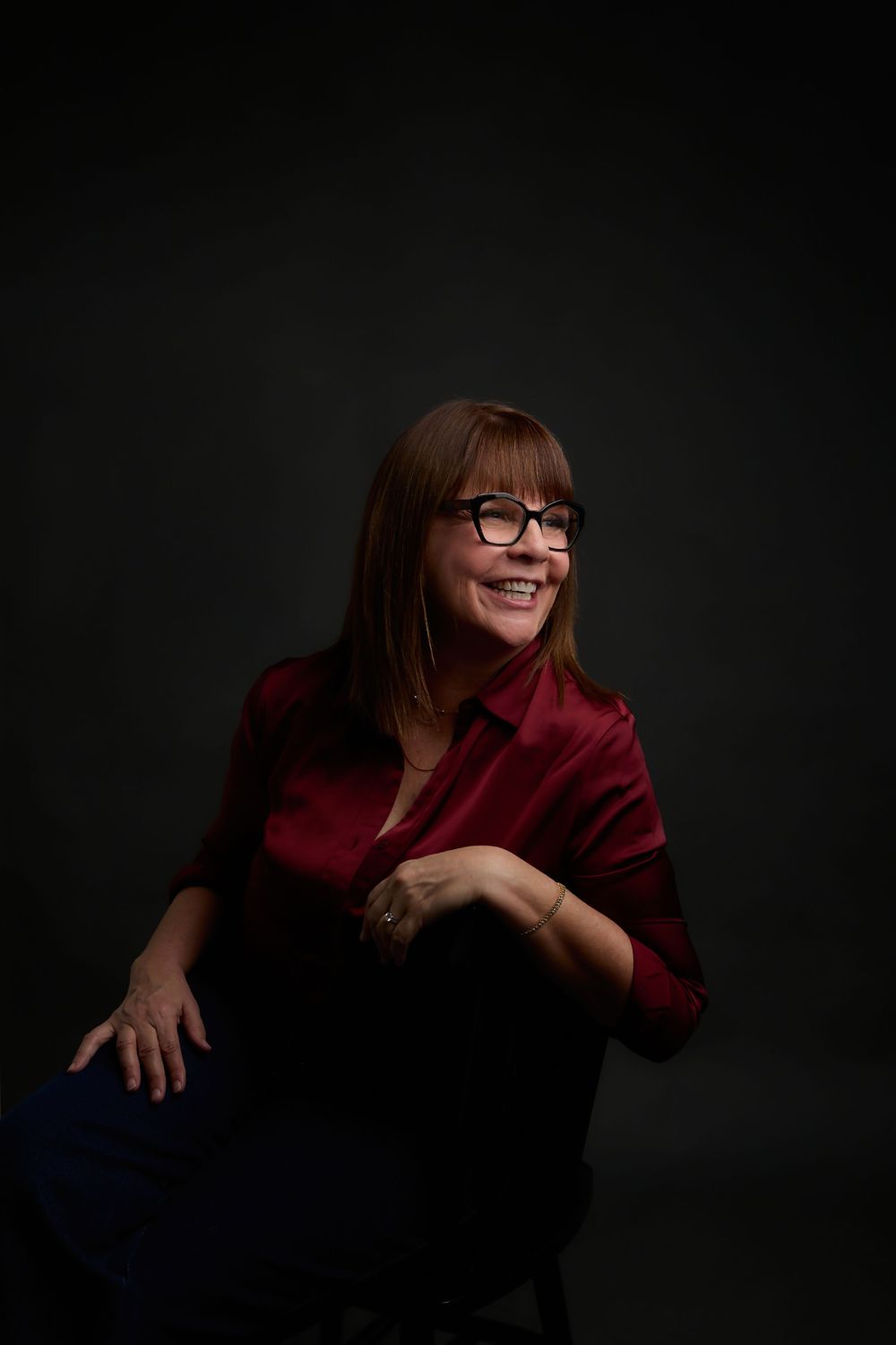 Woman with red hair and glasses smiling, wearing a crimson blazer, seated against a dark studio background.