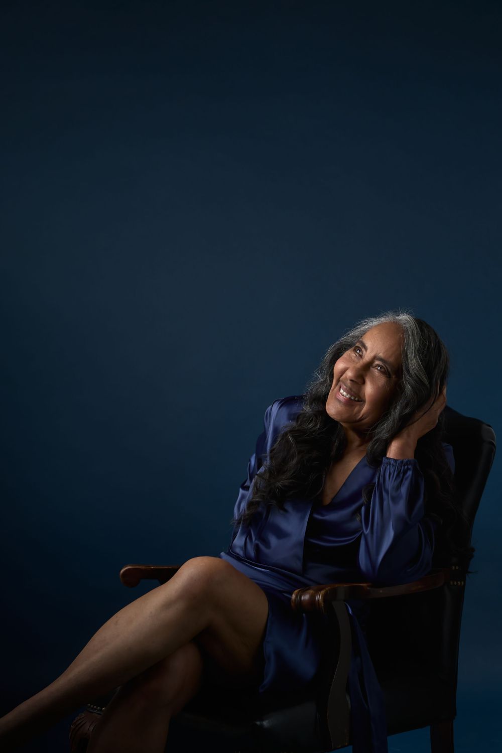 A smiling middle-aged woman with gray-streaked hair sits in a wooden chair wearing a blue satin dress against a dark blue background.
