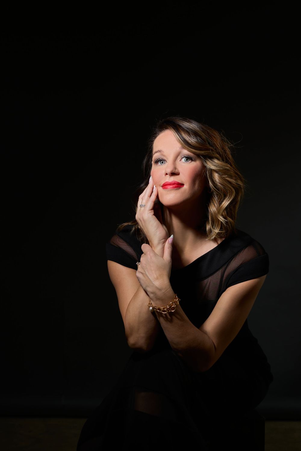 Woman in black dress poses elegantly against dark background, hand on cheek, wearing gold bracelet and red lipstick.