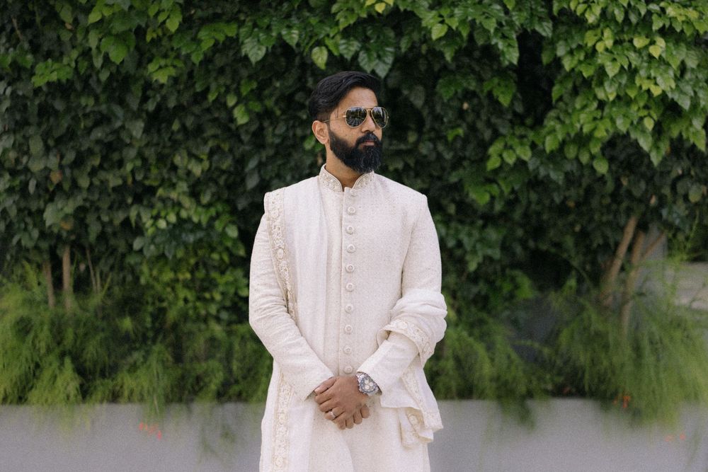 Man in white embroidered sherwani with sunglasses posing outdoors against lush green foliage backdrop.