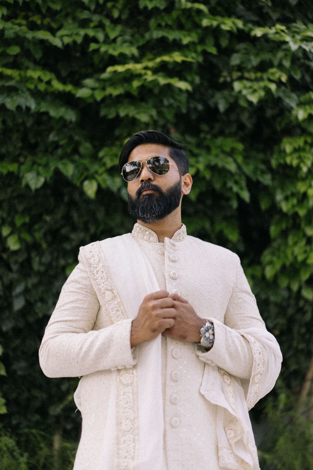 A bearded South Asian man in a cream embroidered sherwani and aviator sunglasses poses outdoors against lush green foliage.