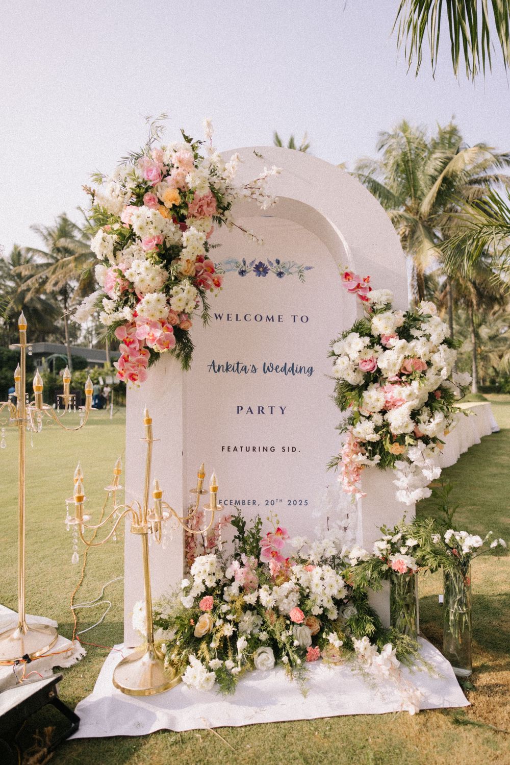 Elegant outdoor wedding welcome sign adorned with pink and white floral arrangements, candelabras, and lush greenery.