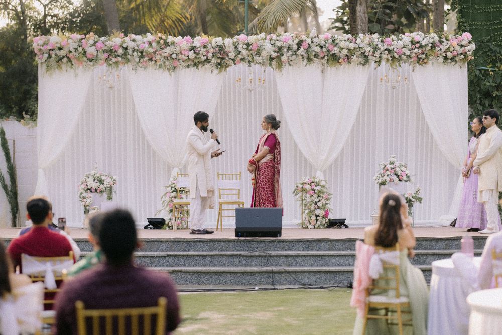 Outdoor Indian wedding ceremony with couple at floral mandap, pink and white flowers, guests seated on lawn.