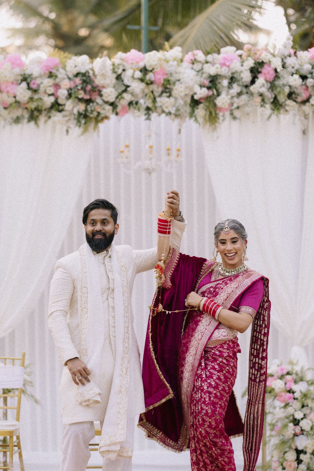 Indian bride in magenta saree and groom in white sherwani celebrate at elegant floral wedding ceremony backdrop.