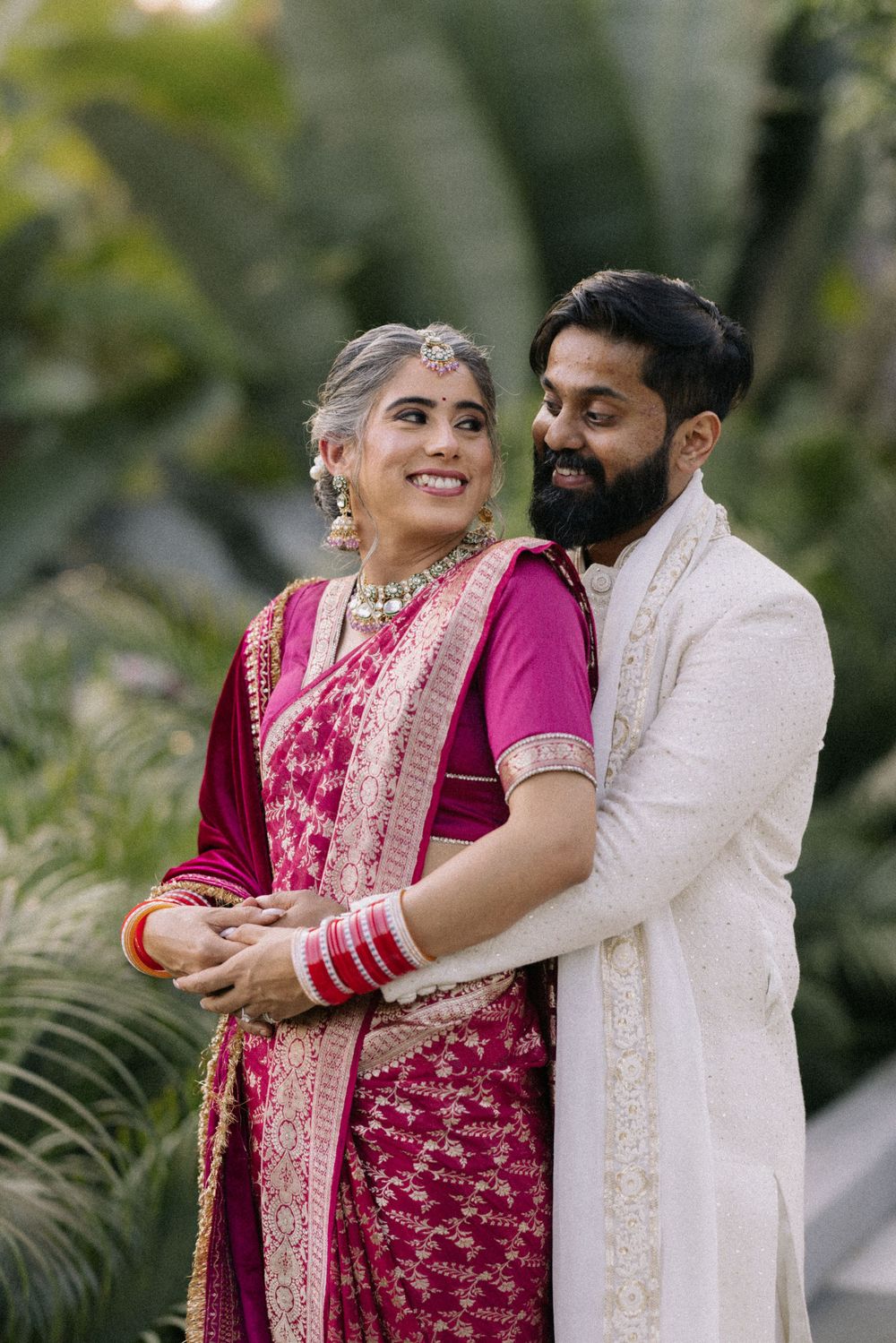 Indian bride in magenta saree and groom in white sherwani embrace outdoors during traditional wedding ceremony.