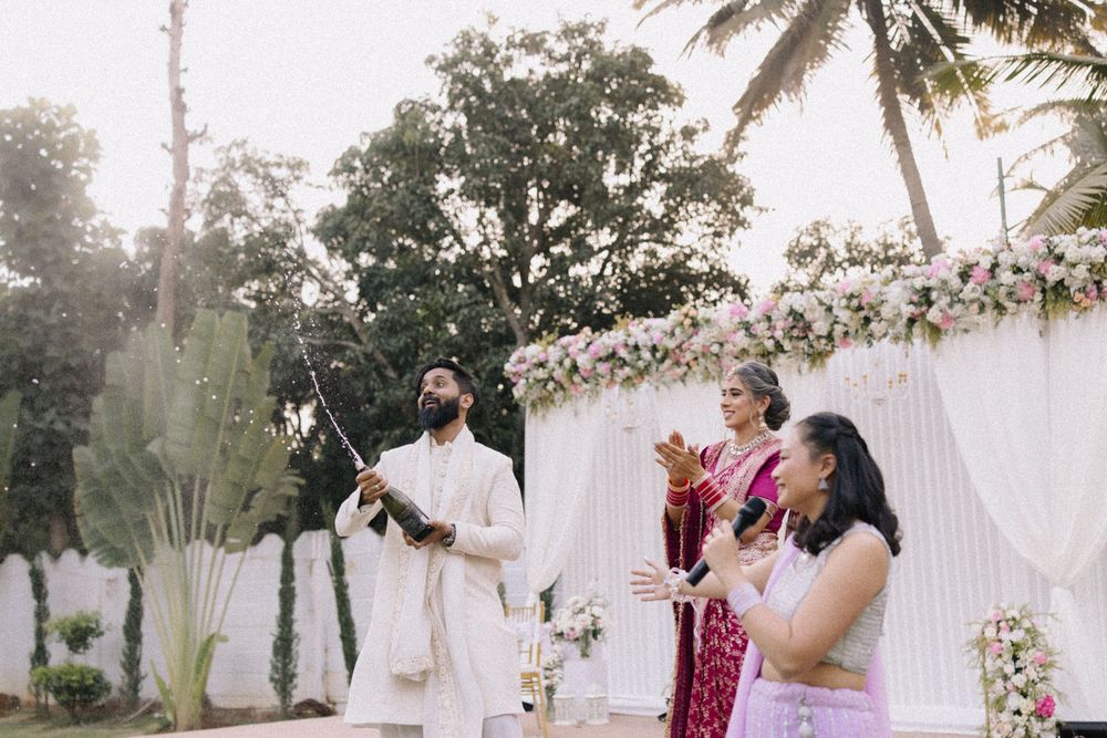 Indian groom in white sherwani performs wedding ritual outdoors with guests near floral arch and palm trees.