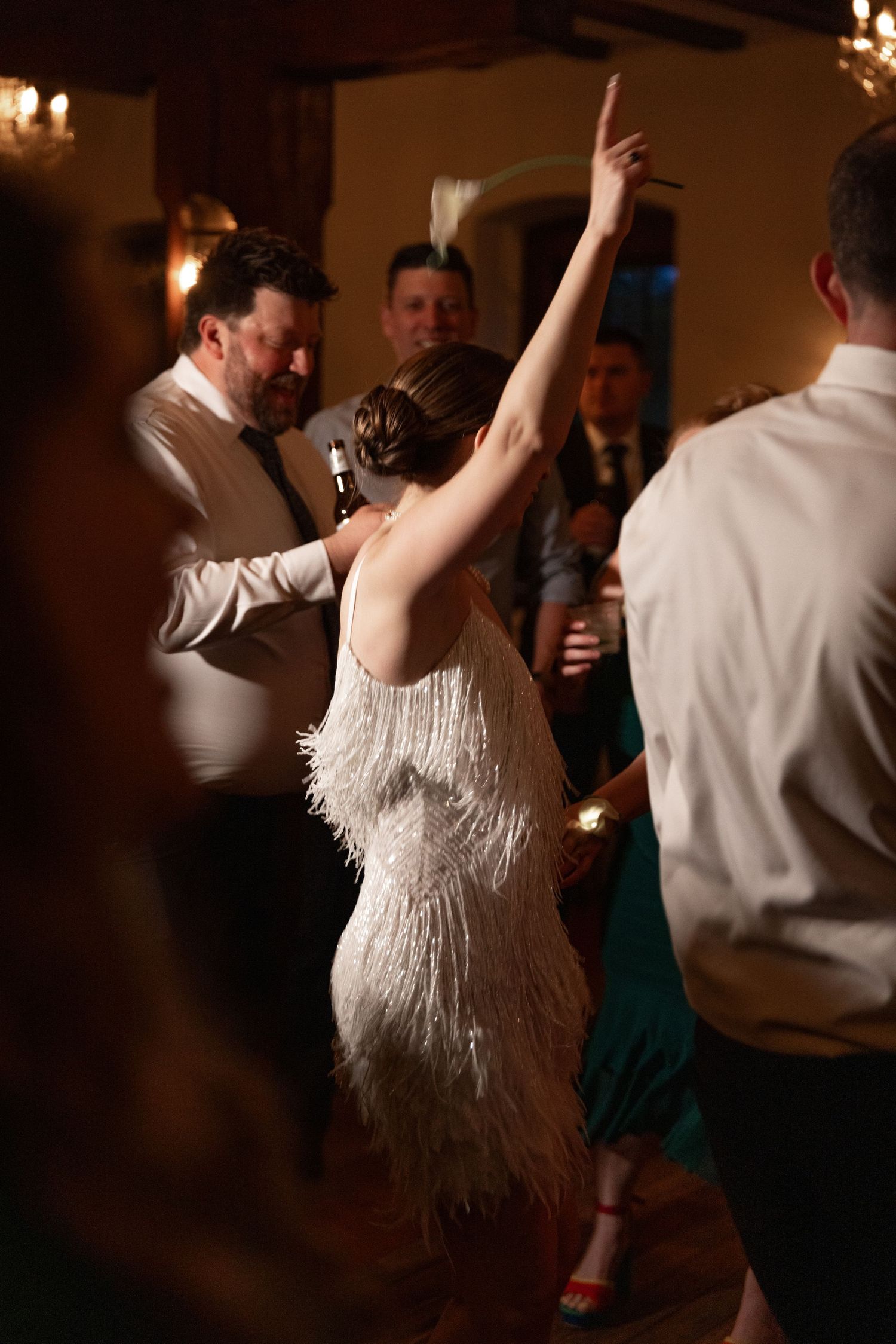 Bride in feathered fringe dress dancing energetically with guests at lively wedding reception.