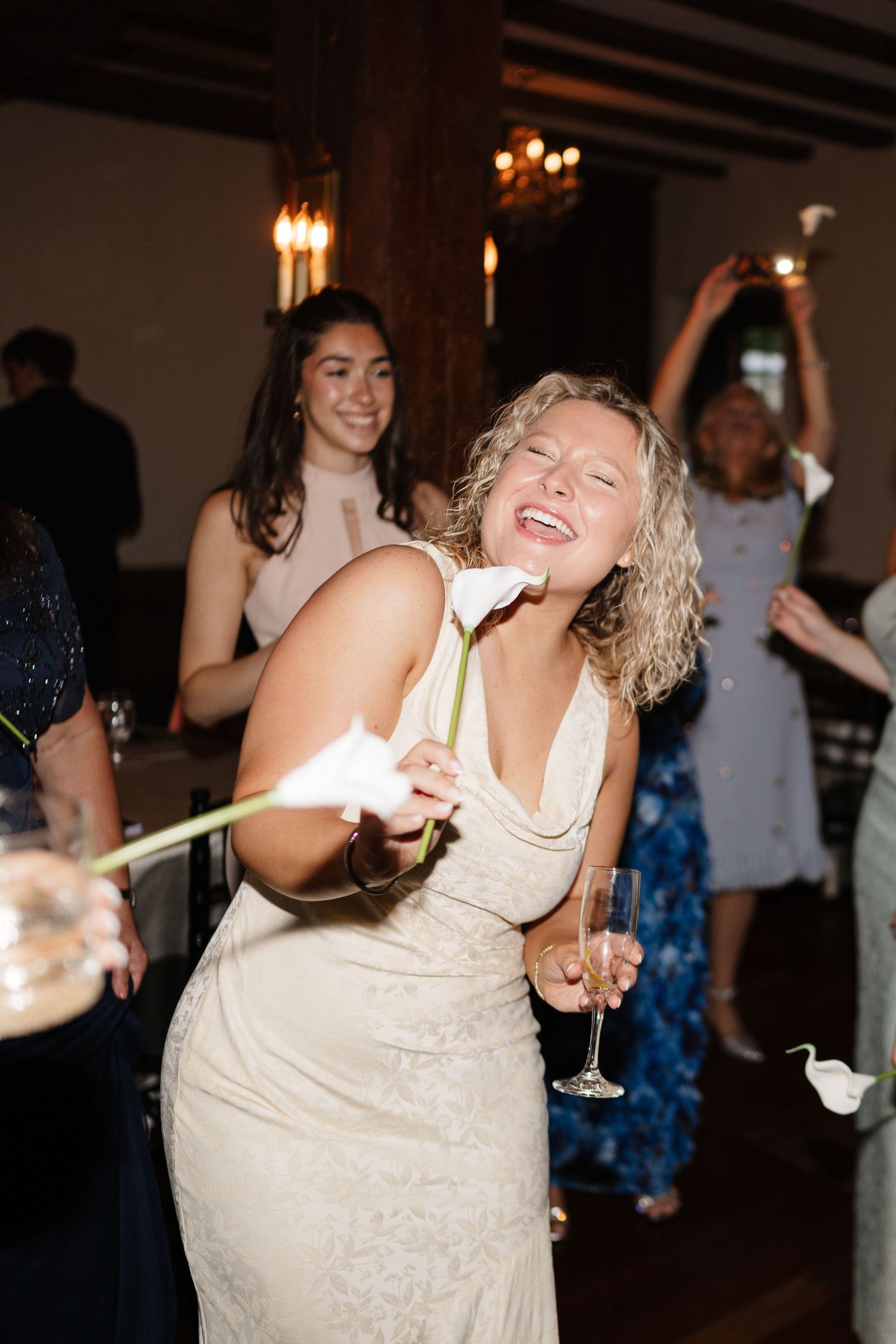 Joyful wedding guest in beige lace dress dancing and holding a white calla lily and champagne glass.