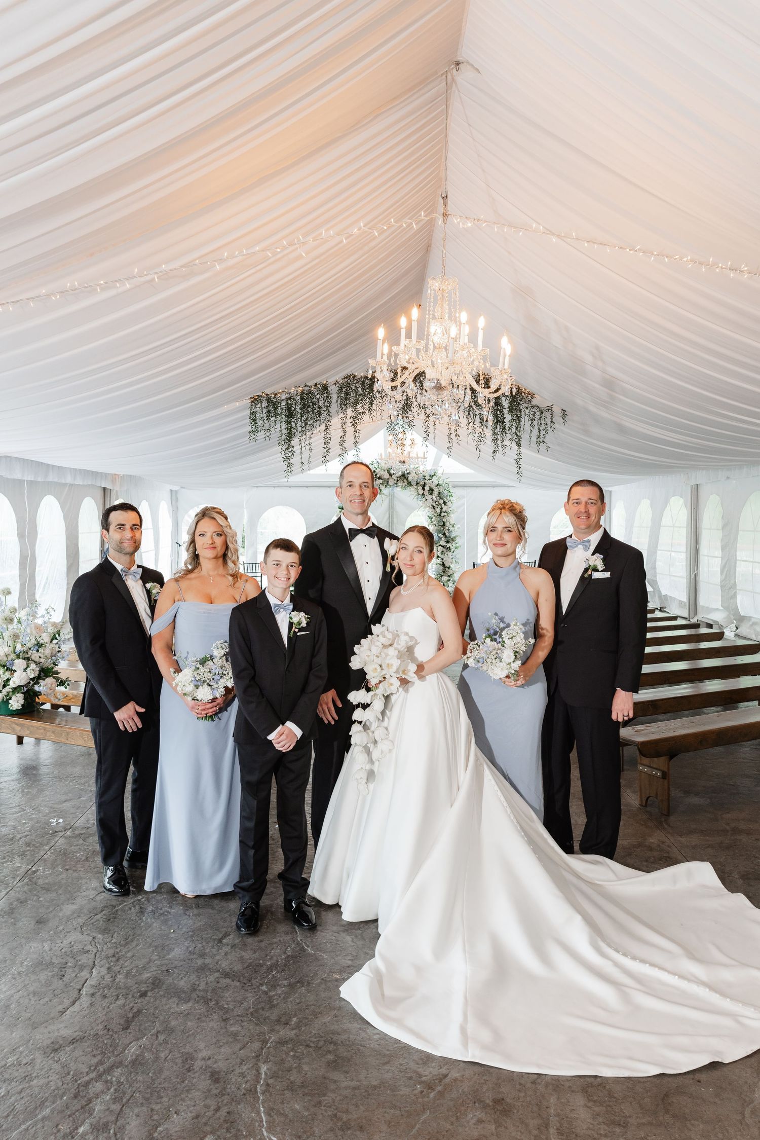 Wedding party poses in elegant white tent with chandelier, bridesmaids in dusty blue gowns flanking bride and groom.