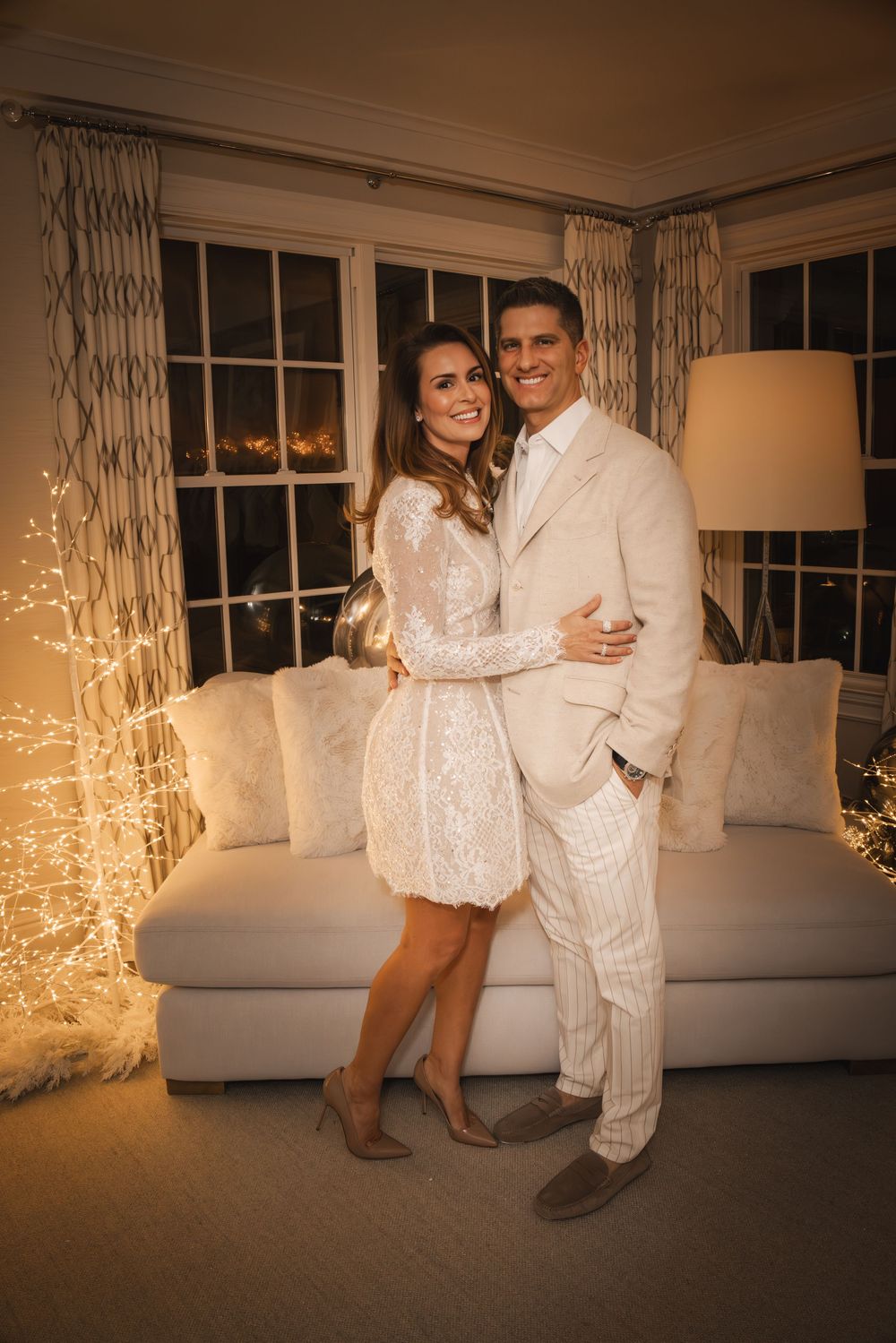 Elegant couple posing in white outfits in a warmly lit living room with fairy lights and a white sofa.