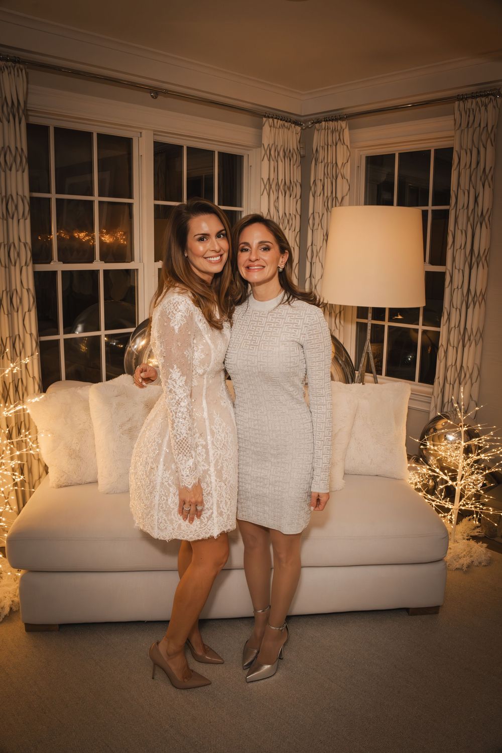 Two women in elegant white dresses pose together in a festively decorated living room with holiday lights.