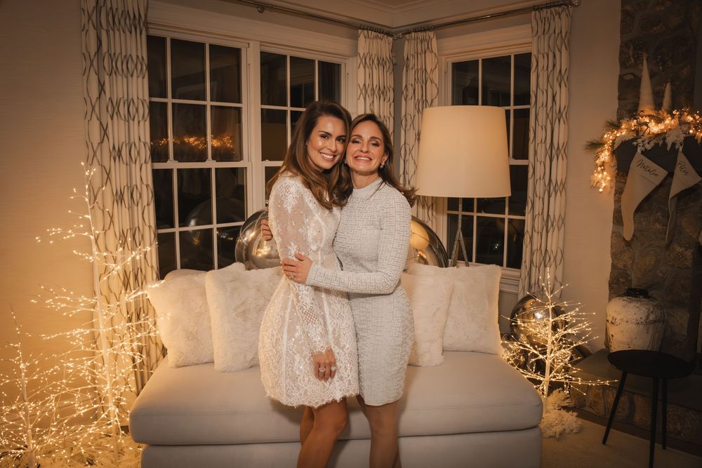 Two women in white dresses pose together in a cozy holiday-decorated living room with warm fairy lights.