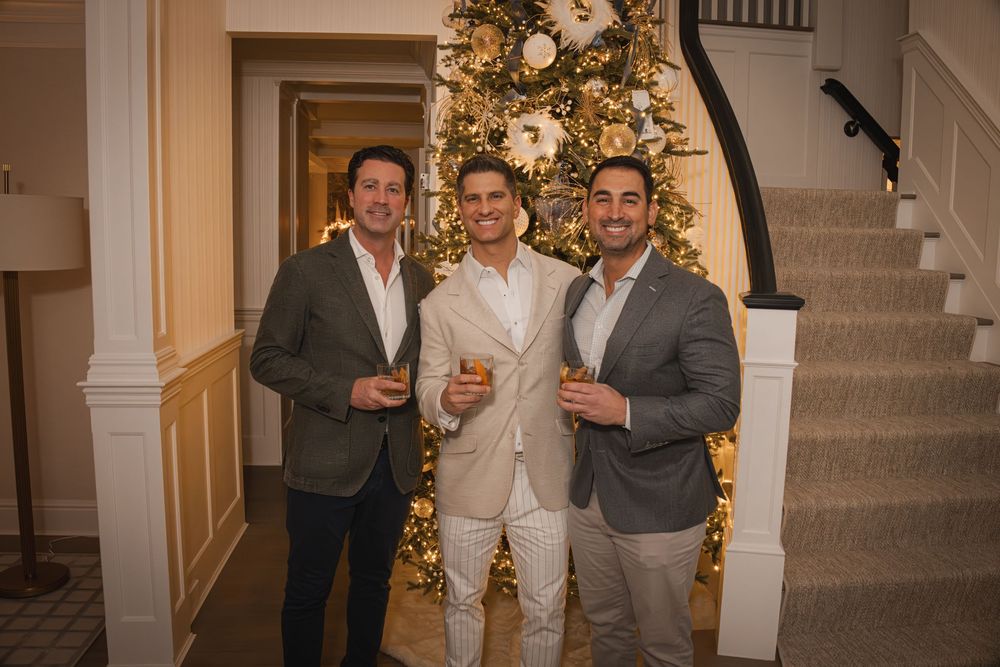 Three men in blazers smile together holding drinks near a decorated Christmas tree at a holiday party.