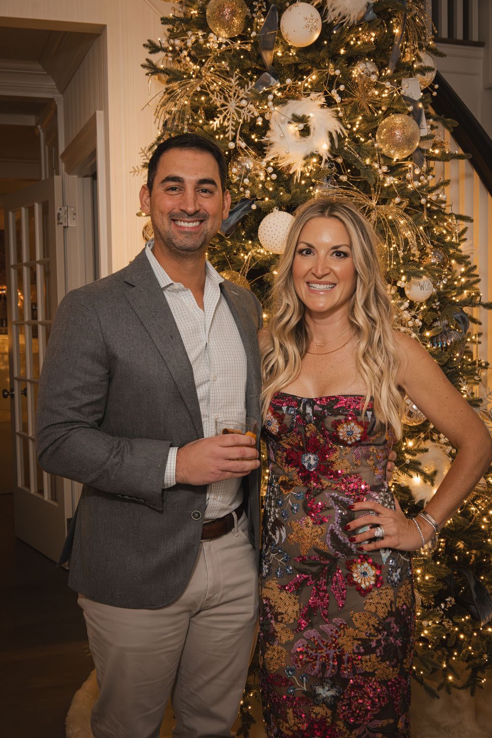 Couple smiling in front of decorated Christmas tree at holiday party, man in gray blazer, woman in floral dress.
