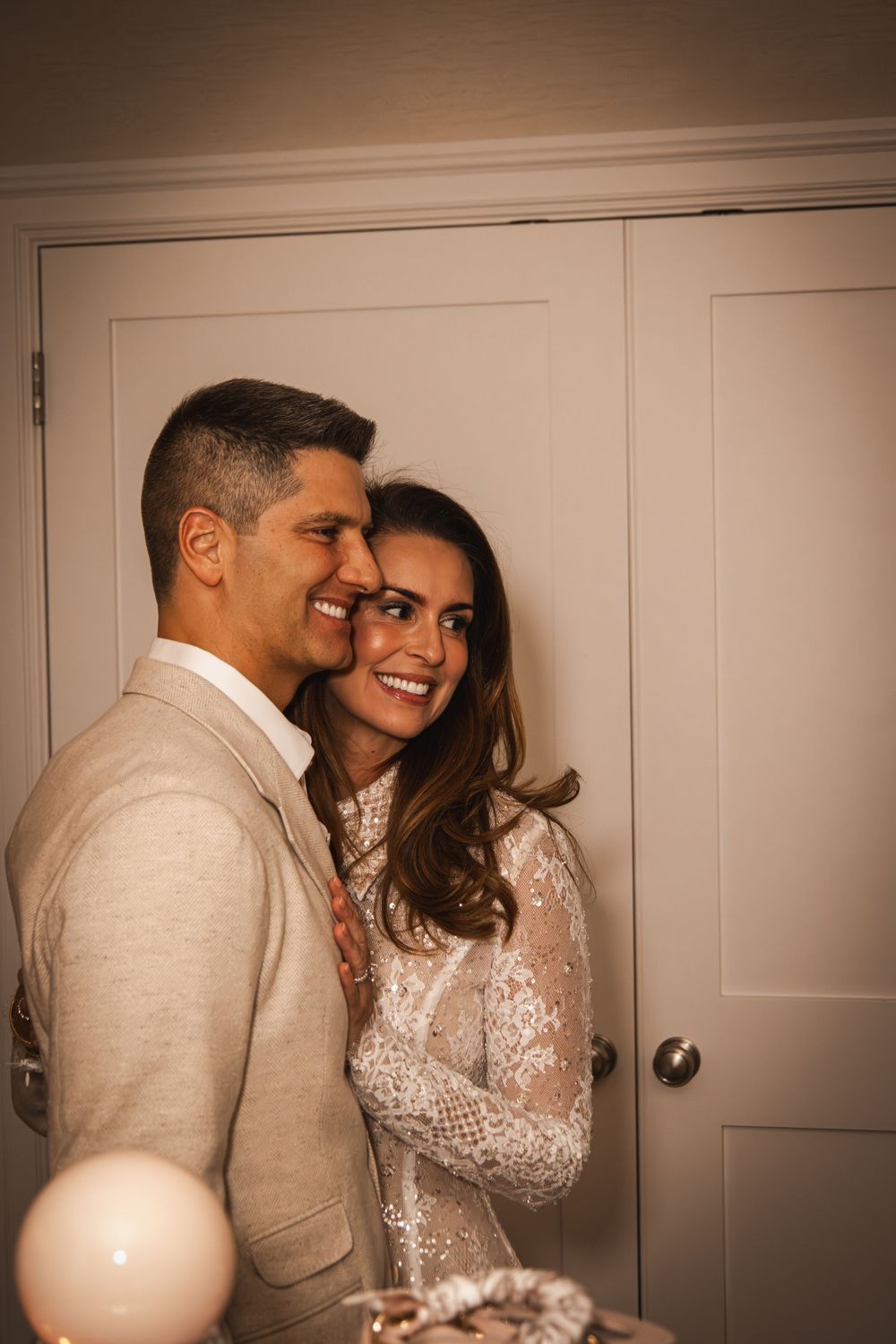 A smiling couple in elegant attire embraces warmly in a softly lit room, suggesting a wedding or special celebration.