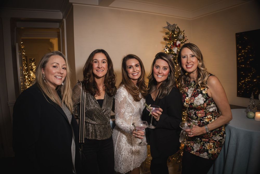 Five women smiling at a festive holiday party with a decorated Christmas tree in the background.