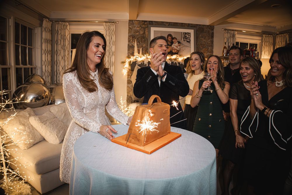 Woman in white lace dress smiles beside sparkler-lit cake at elegant indoor celebration with guests.