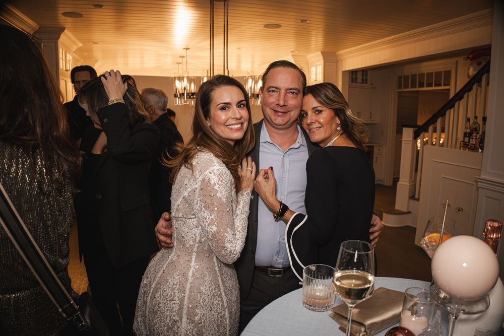 Three friends smiling and posing together at an elegant indoor party with warm lighting and champagne glasses.