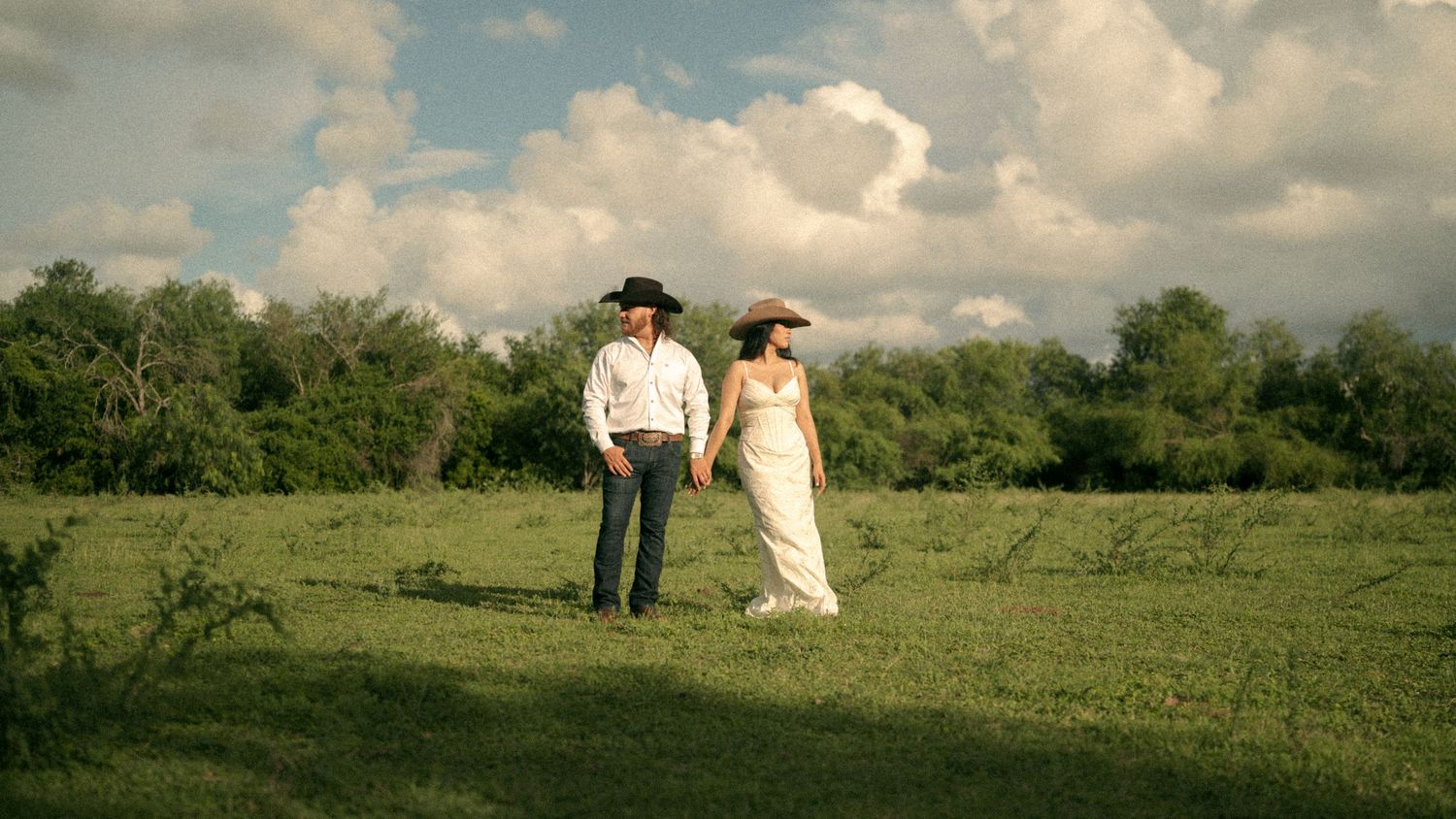 Couple in cowboy hats holding hands walking through a green meadow under a cloudy sky in western wedding attire.