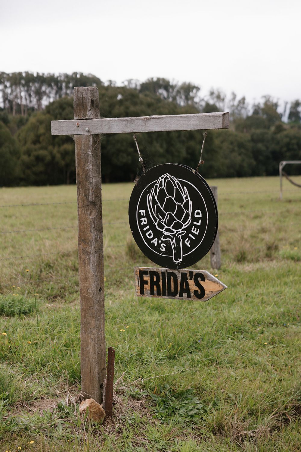 Wooden directional sign post with black circular logo pointing to Frida's restaurant in a grassy field.