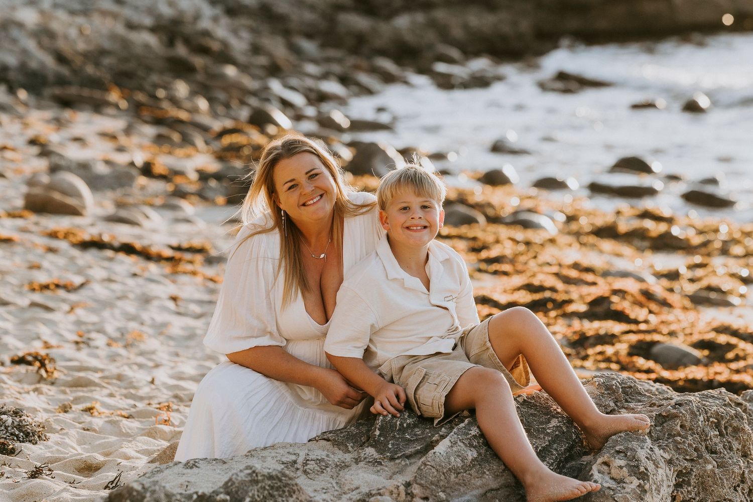 Family of four smiling together on rocky beach at golden sunset with ocean backdrop.