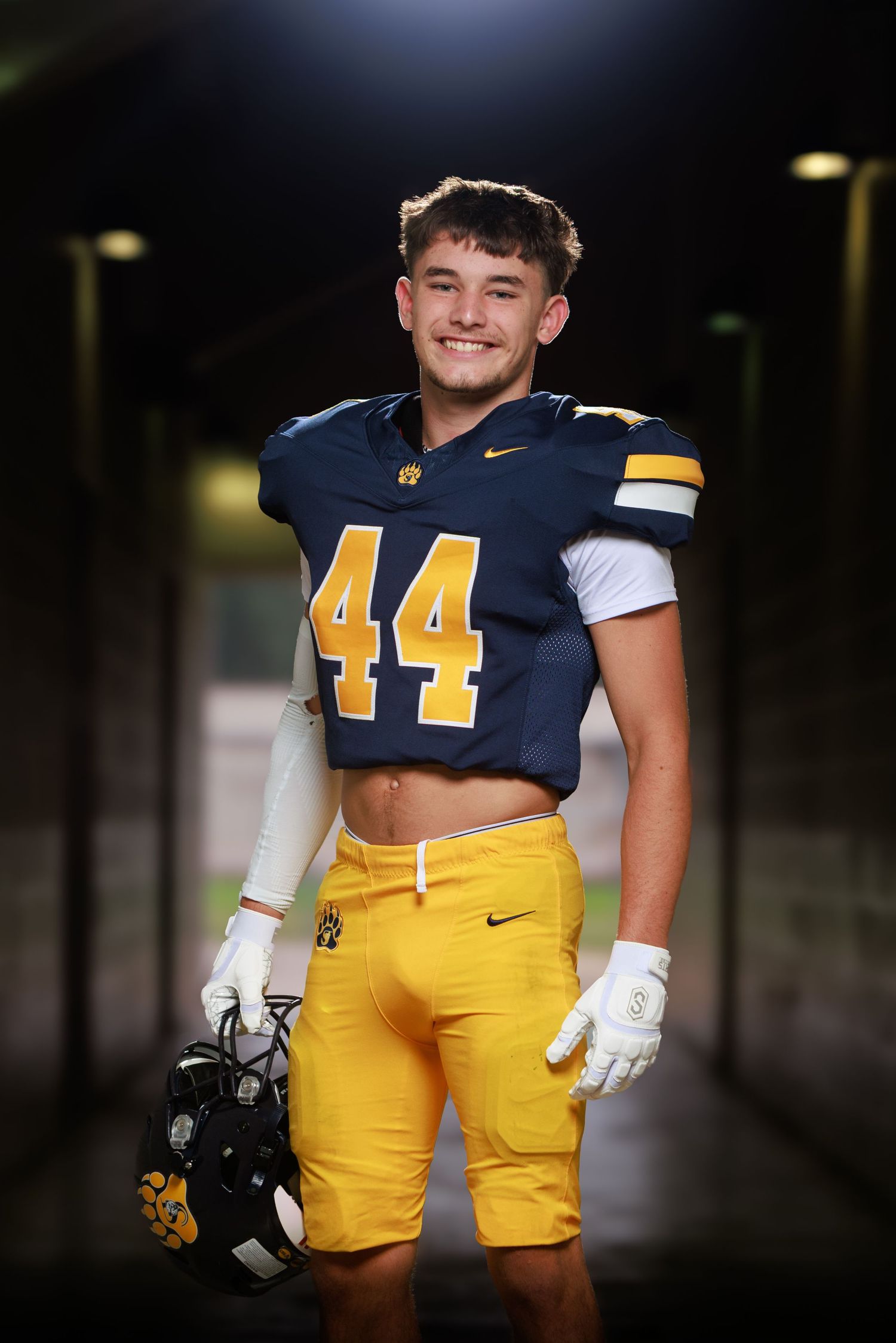 Football player #12 in navy and gold uniform poses at Birchfield Stadium with helmet and ball.