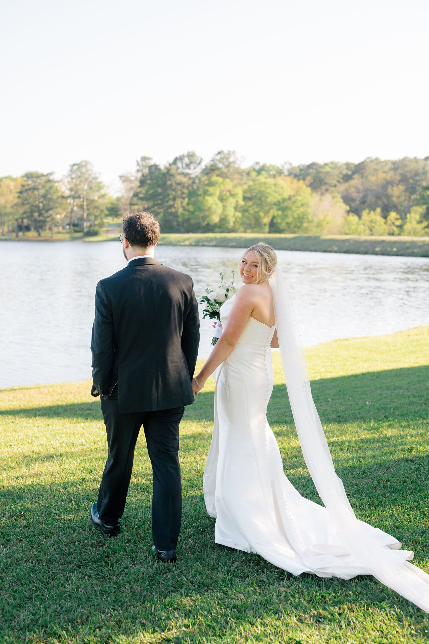Bride in white gown with veil and groom in black suit hold hands by a serene lakeside on their wedding day.