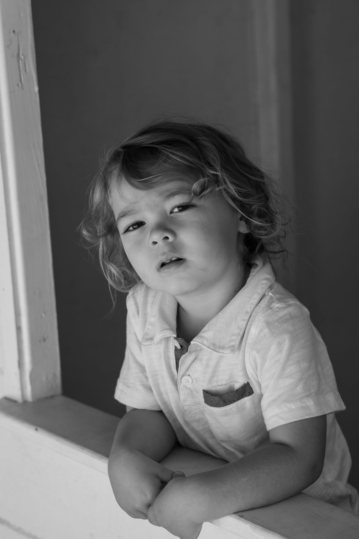 Black and white portrait of a pensive young child with curly hair leaning on a windowsill indoors.