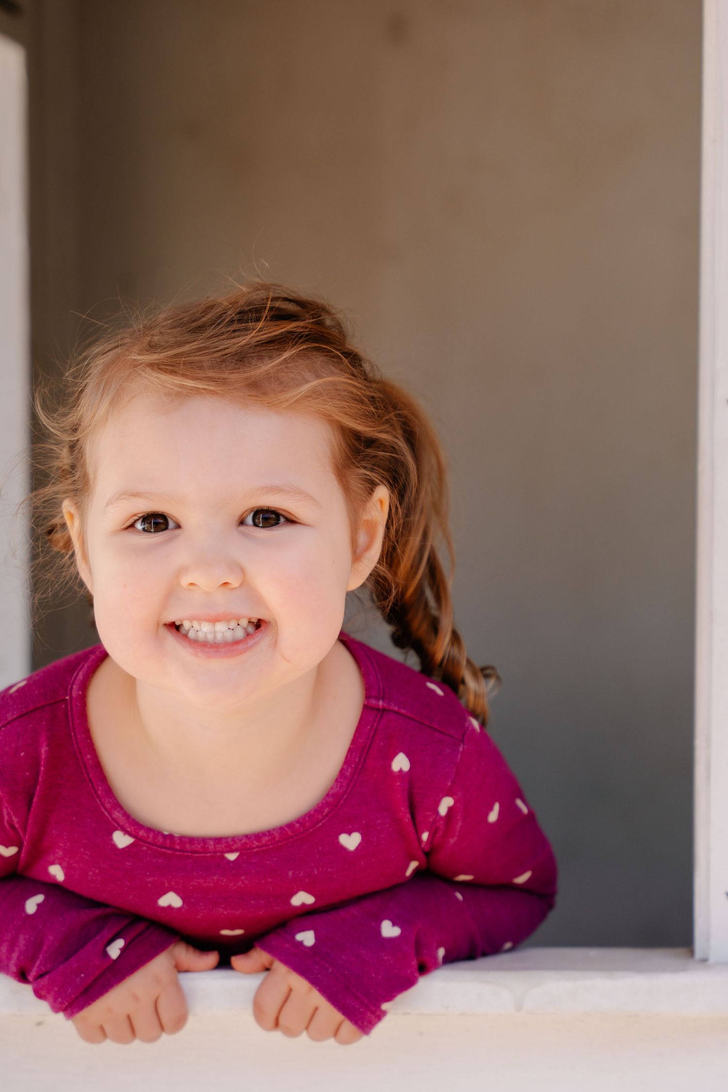 Smiling toddler girl with braids wearing a pink heart-print sweater, leaning on a white ledge.