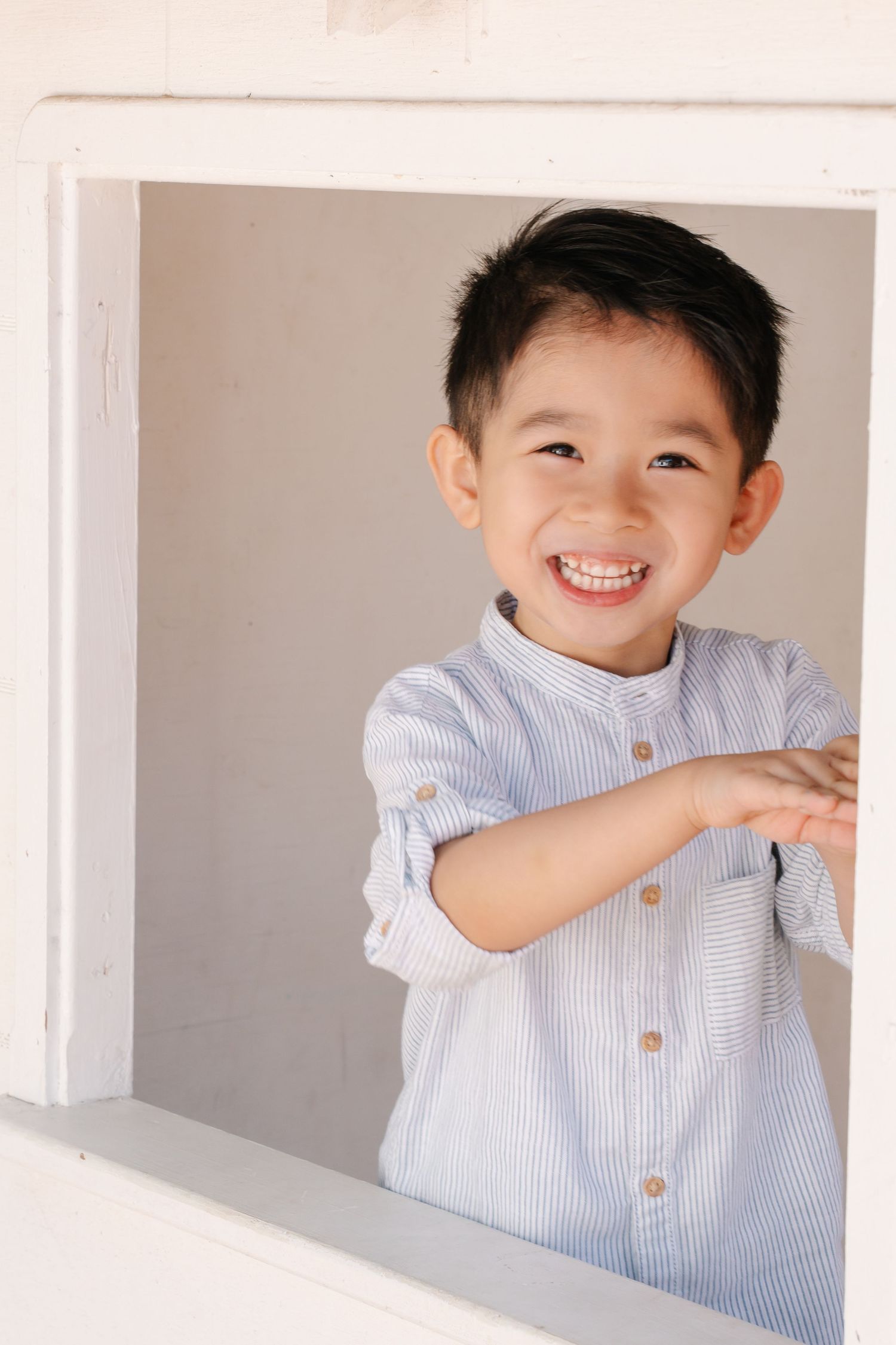 A smiling young Asian boy in a blue striped shirt leans out of a white window frame, looking cheerful and playful.