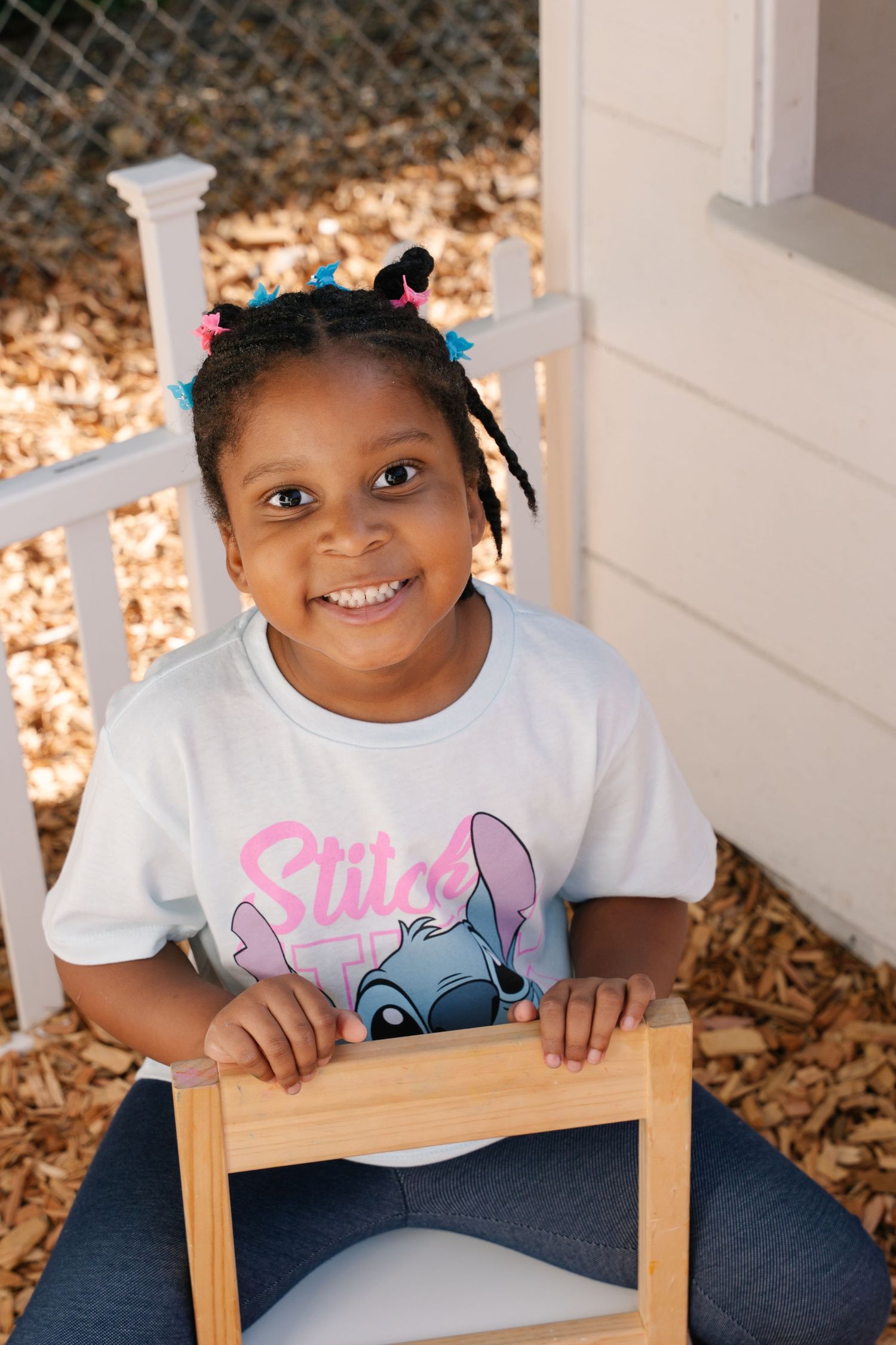 Happy young girl in Stitch t-shirt with colorful braids sitting on chair outdoors.
