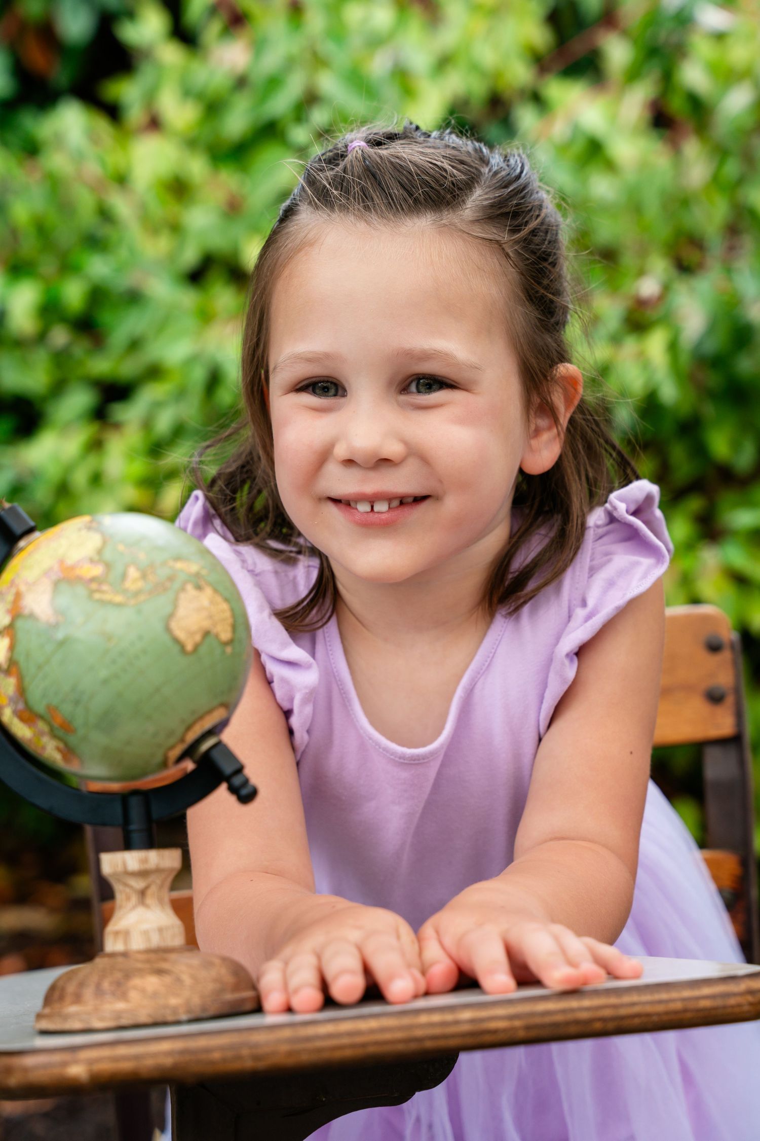 Happy little girl in purple dress leaning on desk with vintage globe in green garden setting.